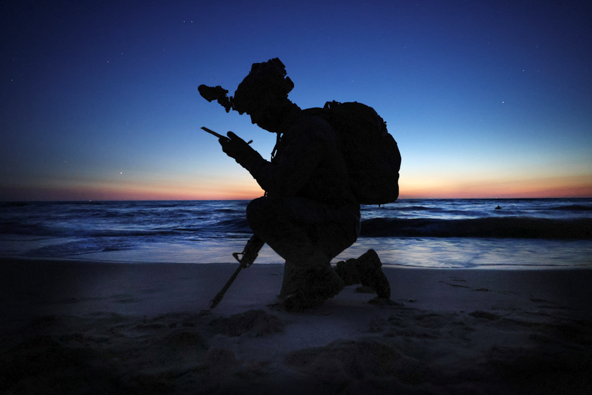 A soldier in silhouette uses a radio on a beach