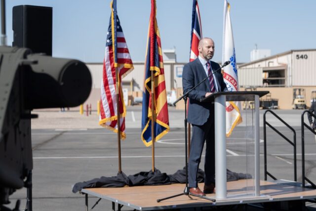 DE&S Director Rotary Wing & UAS, Mark Langrill, speaking in front of flags on poles at the acceptance of the 50th and final AH-64E Apache attack helicopter at Boeing's manufacturing facility in Arizona