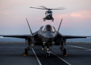 An F-35B Lightning II fighter jet on the deck of HMS Prince of Wales, with an airborne Merlin helicopter behind it during the Royal Navy's Carrier Strike Group deployment in 2025