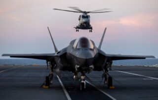 An F-35B Lightning II fighter jet on the deck of HMS Prince of Wales, with an airborne Merlin helicopter behind it during the Royal Navy's Carrier Strike Group deployment in 2025