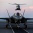 An F-35B Lightning II fighter jet on the deck of HMS Prince of Wales, with an airborne Merlin helicopter behind it during the Royal Navy's Carrier Strike Group deployment in 2025