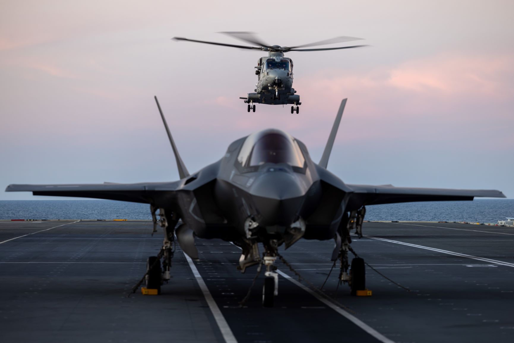 An F-35B Lightning II fighter jet on the deck of HMS Prince of Wales, with an airborne Merlin helicopter behind it during the Royal Navy's Carrier Strike Group deployment in 2025